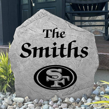 Custom San Francisco 49ers Name Stone displayed in a landscaped flower bed, showcasing personalized family lettering and officially licensed team artwork. This decorative outdoor stone creates a striking focal point for 49ers fans and outdoor living spaces.