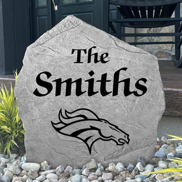 Custom Denver Broncos Name Stone displayed in a landscaped flower bed, showcasing personalized family lettering and officially licensed team artwork. This decorative outdoor stone creates a striking focal point for Broncos fans and outdoor living spaces.