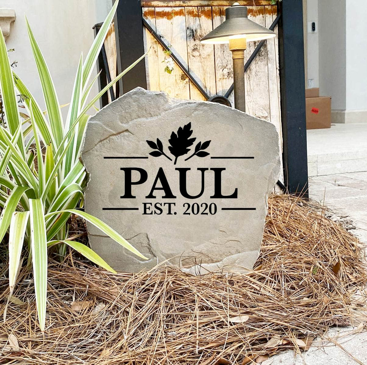 Personalized Fall Name Stone reading PAUL Est. 2020 on pine straw beside a rustic garden gate and landscape light