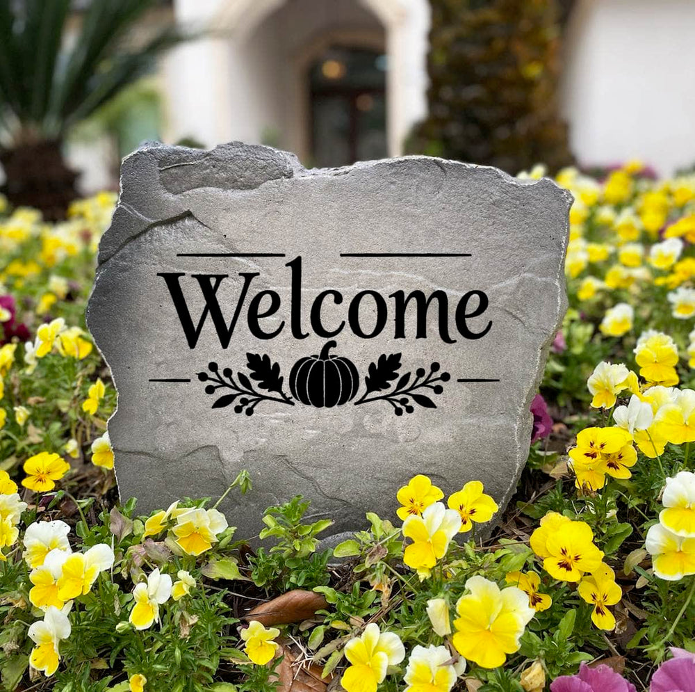Gray Fall Welcome Stone standing in a bed of yellow and purple pansies, engraved Welcome with a pumpkin design
