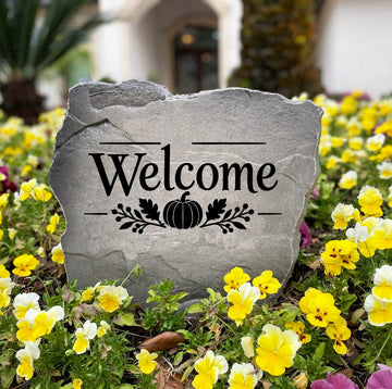 Gray Fall Welcome Stone standing in a bed of yellow and purple pansies, engraved Welcome with a pumpkin design