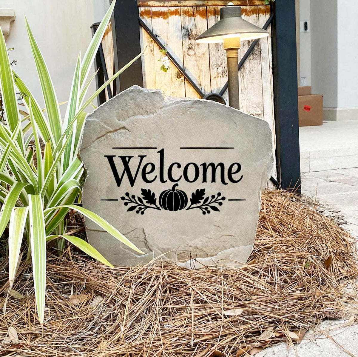 Fall Welcome Stone on pine straw next to a rustic garden gate and landscape light, engraved with a pumpkin motif