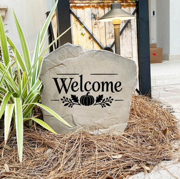 Fall Welcome Stone on pine straw next to a rustic garden gate and landscape light, engraved with a pumpkin motif