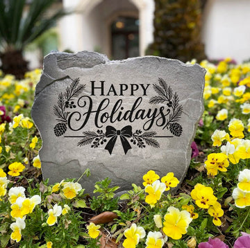 Gray Happy Holidays Stone standing in a bed of yellow and purple pansies, engraved with pine and a bow
