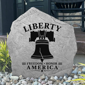 Decorative stone with 'Liberty Freedom Honor America' text and bell design on a pebble-covered ground.