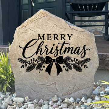 Sandstone Merry Christmas Stone engraved with script lettering, pine sprigs and a bow, set in river rock