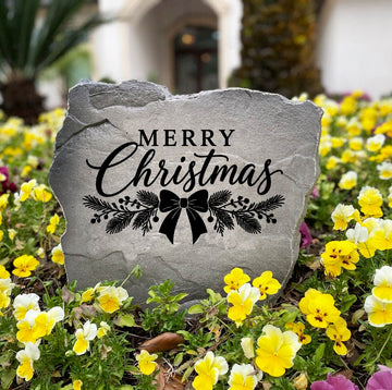 Gray Merry Christmas Stone standing in a bed of yellow and purple pansies, engraved with a ribbon bow