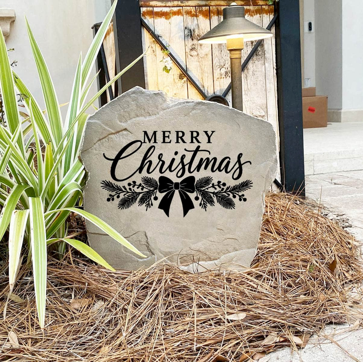Merry Christmas Stone on pine straw next to a rustic garden gate and landscape light