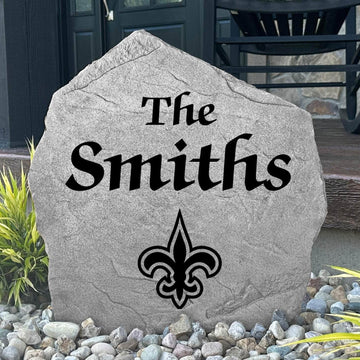Custom New Orleans Saints Name Stone displayed in a landscaped flower bed, showcasing personalized family lettering and officially licensed team artwork. This decorative outdoor stone creates a striking focal point for Saints fans and outdoor living spaces.