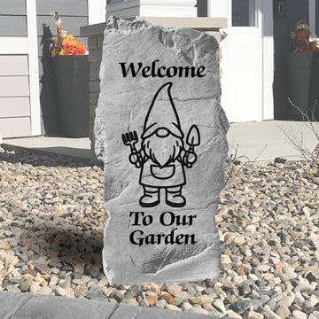 Decorative stone with a garden gnome design and 'Welcome To Our Garden' text on a pebbled outdoor setting.