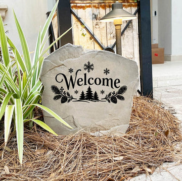 Winter Welcome Stone on pine straw next to a rustic garden gate and landscape light, engraved with pine trees