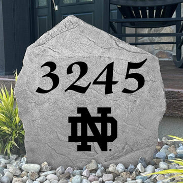 Custom University of Notre Dame Address Stone displayed in a landscaped flower bed, showcasing personalized house numbers and officially licensed Fighting Irish artwork. This engraved address stone creates a beautiful focal point for alumni homes and outdoor spaces.