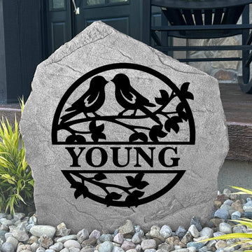 Custom Love Birds Monogram Stone displayed in a landscaped flower bed, showcasing romantic bird artwork and personalized engraving. This engraved outdoor stone creates a beautiful focal point for walkways and home entrances.