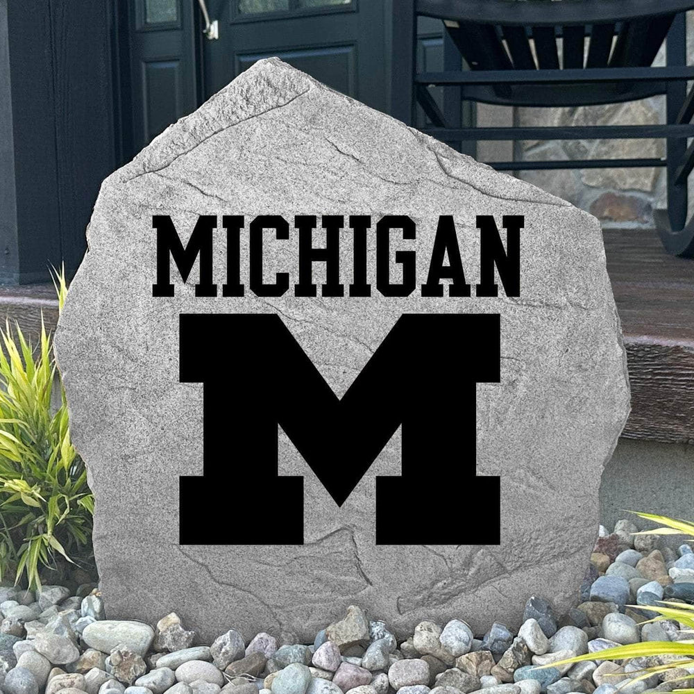 Custom University of Michigan Logo Stone displayed in a landscaped flower bed, showcasing officially licensed Wolverines artwork with permanent engraving. This decorative outdoor stone creates a beautiful focal point for alumni homes and Michigan fans.