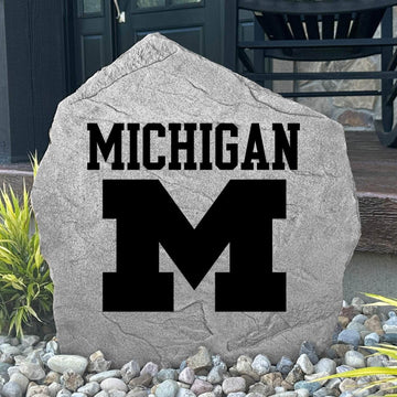 Custom University of Michigan Logo Stone displayed in a landscaped flower bed, showcasing officially licensed Wolverines artwork with permanent engraving. This decorative outdoor stone creates a beautiful focal point for alumni homes and Michigan fans.