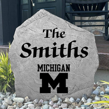 Custom University of Michigan Name Stone displayed in a landscaped flower bed, showcasing personalized family lettering and officially licensed Wolverines artwork. This engraved outdoor stone creates a beautiful focal point for alumni homes and Michigan fans.