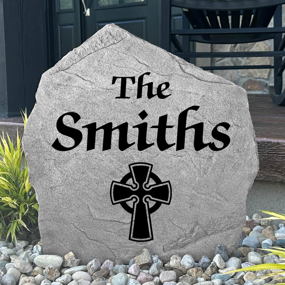 Custom Celtic Cross Name Stone displayed in a landscaped flower bed, showcasing personalized family names and intricate Celtic detailing. This engraved outdoor stone creates a meaningful focal point for walkways and home entrances.