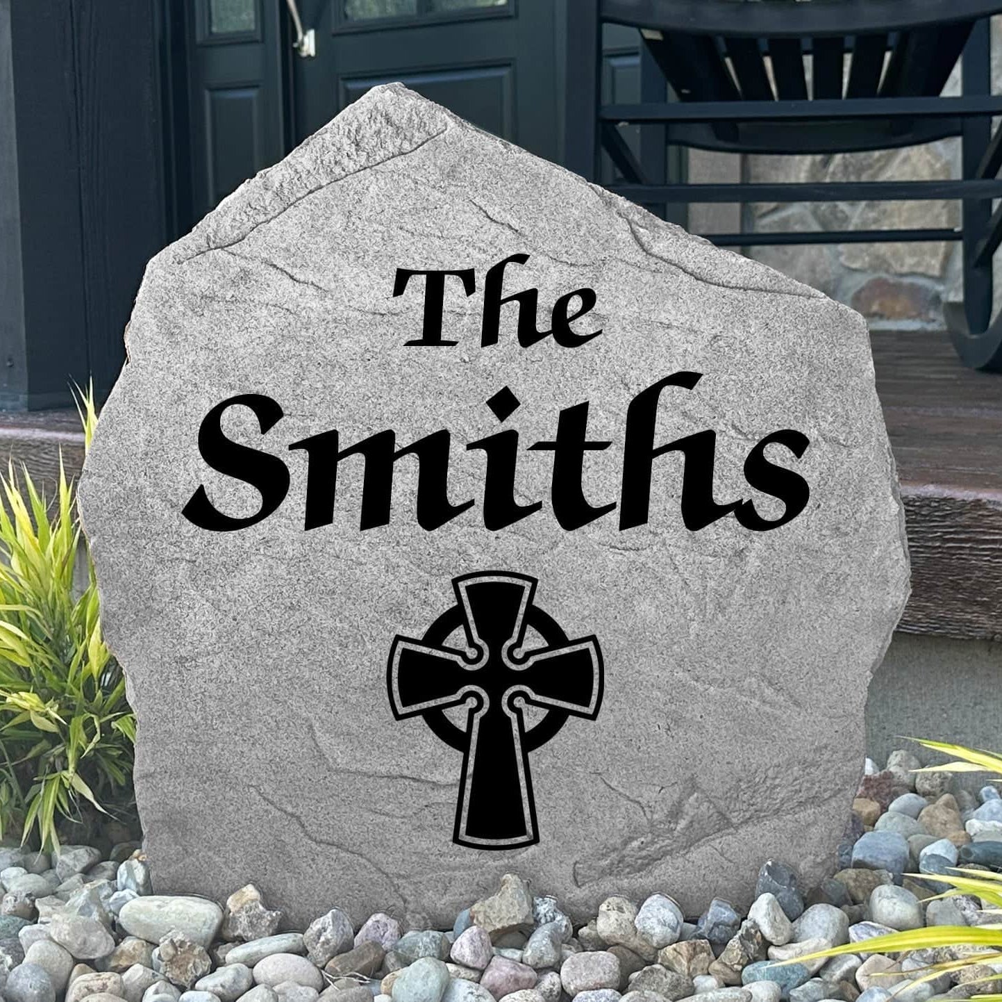 Custom Celtic Cross Name Stone displayed in a landscaped flower bed, showcasing personalized family names and intricate Celtic detailing. This engraved outdoor stone creates a meaningful focal point for walkways and home entrances.