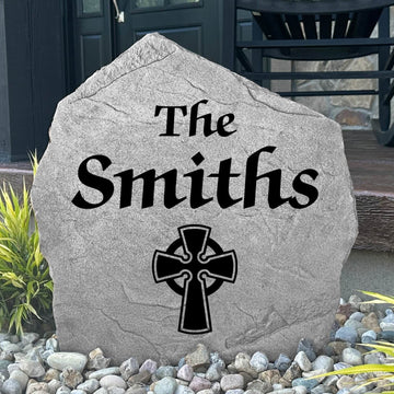 Custom Celtic Cross Name Stone displayed in a landscaped flower bed, showcasing personalized family names and intricate Celtic detailing. This engraved outdoor stone creates a meaningful focal point for walkways and home entrances.