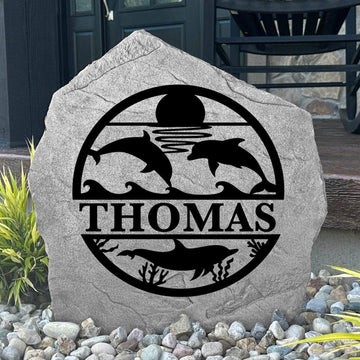 Custom Dolphin Monogram Stone displayed in a landscaped flower bed, showcasing personalized engraving and ocean-inspired dolphin artwork. This engraved outdoor stone creates a beautiful focal point for walkways and coastal homes.