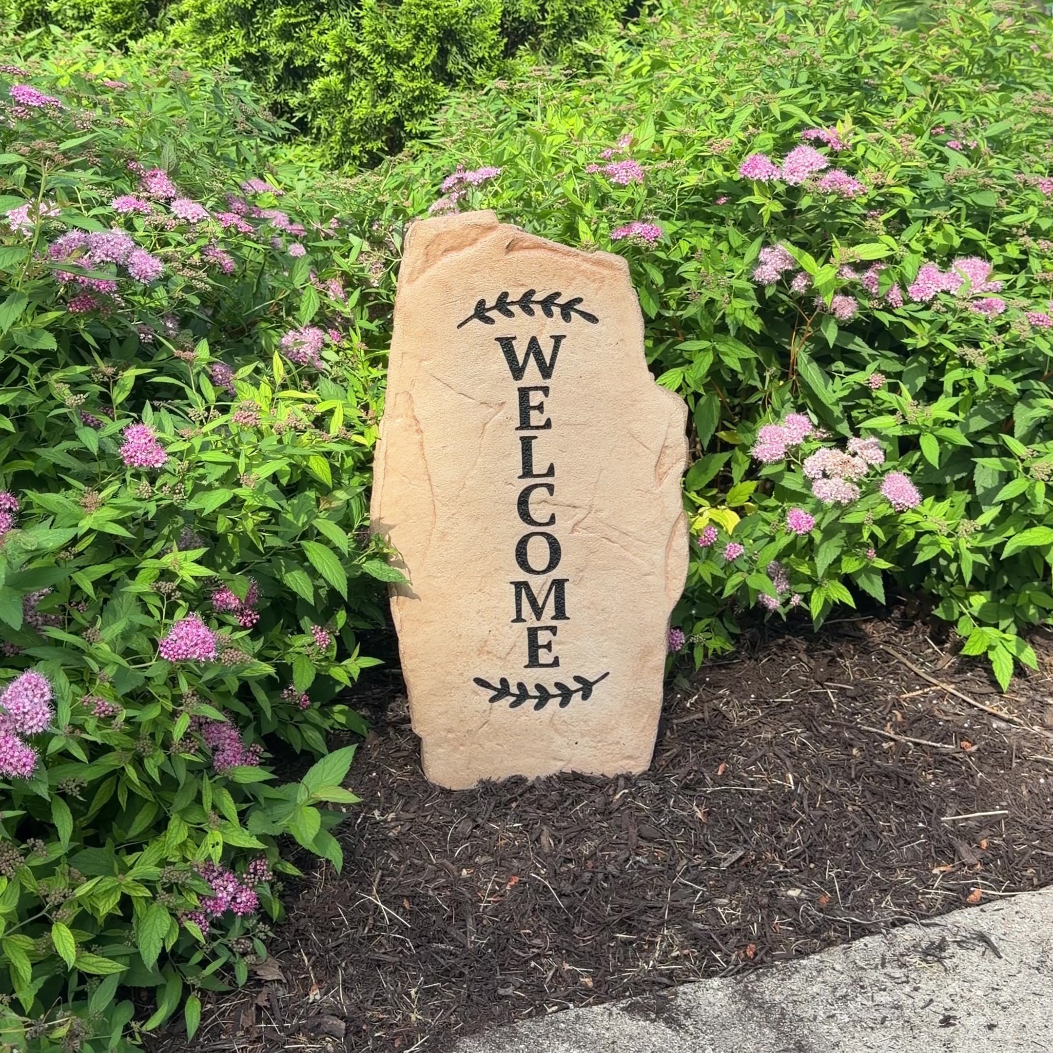 Standing tall beside a front porch entry, the Vertical Welcome Stone offers a stylish greeting with its sleek upright design and beautifully engraved lettering. Framed by colorful flowers and decorative greenery, it creates a warm first impression while adding elegant charm to narrow spaces and landscape beds.
