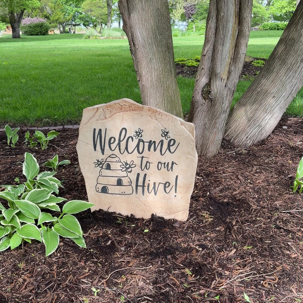Engraved Welcome Hive Stone nestled among colorful flowers and greenery, bringing whimsical charm and nature-inspired beauty to outdoor living spaces. This weather-resistant garden stone creates a warm and inviting entrance.