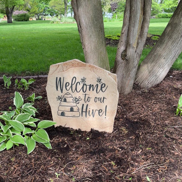 Engraved Welcome Hive Stone nestled among colorful flowers and greenery, bringing whimsical charm and nature-inspired beauty to outdoor living spaces. This weather-resistant garden stone creates a warm and inviting entrance.