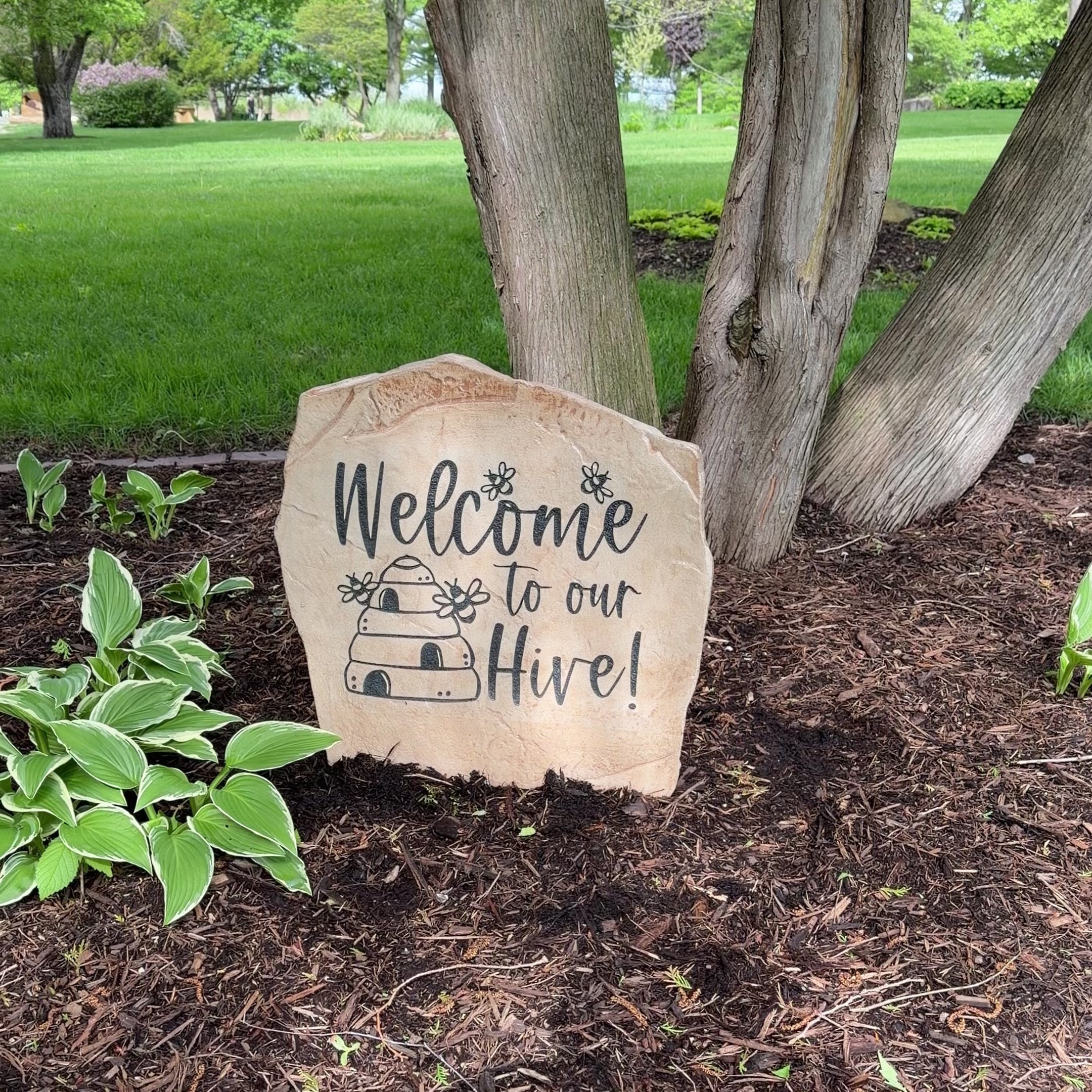 Engraved Welcome Hive Stone nestled among colorful flowers and greenery, bringing whimsical charm and nature-inspired beauty to outdoor living spaces. This weather-resistant garden stone creates a warm and inviting entrance.