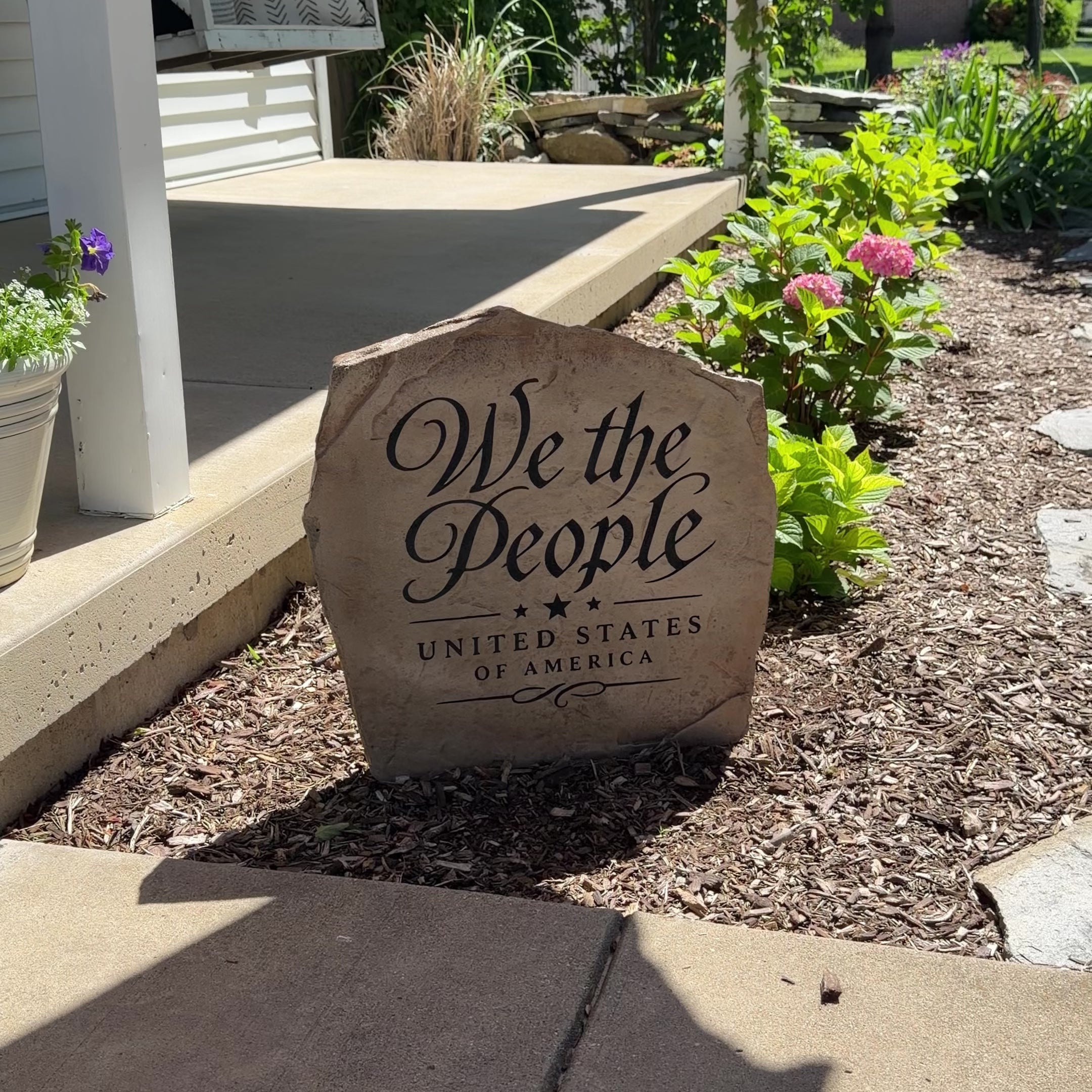 Framed by vibrant landscaping and natural stone, the We The People Stone features elegant engraved script inspired by America's founding principles. Its refined design creates a timeless statement piece that brings patriotic character and classic beauty to a front yard, garden, or entryway.