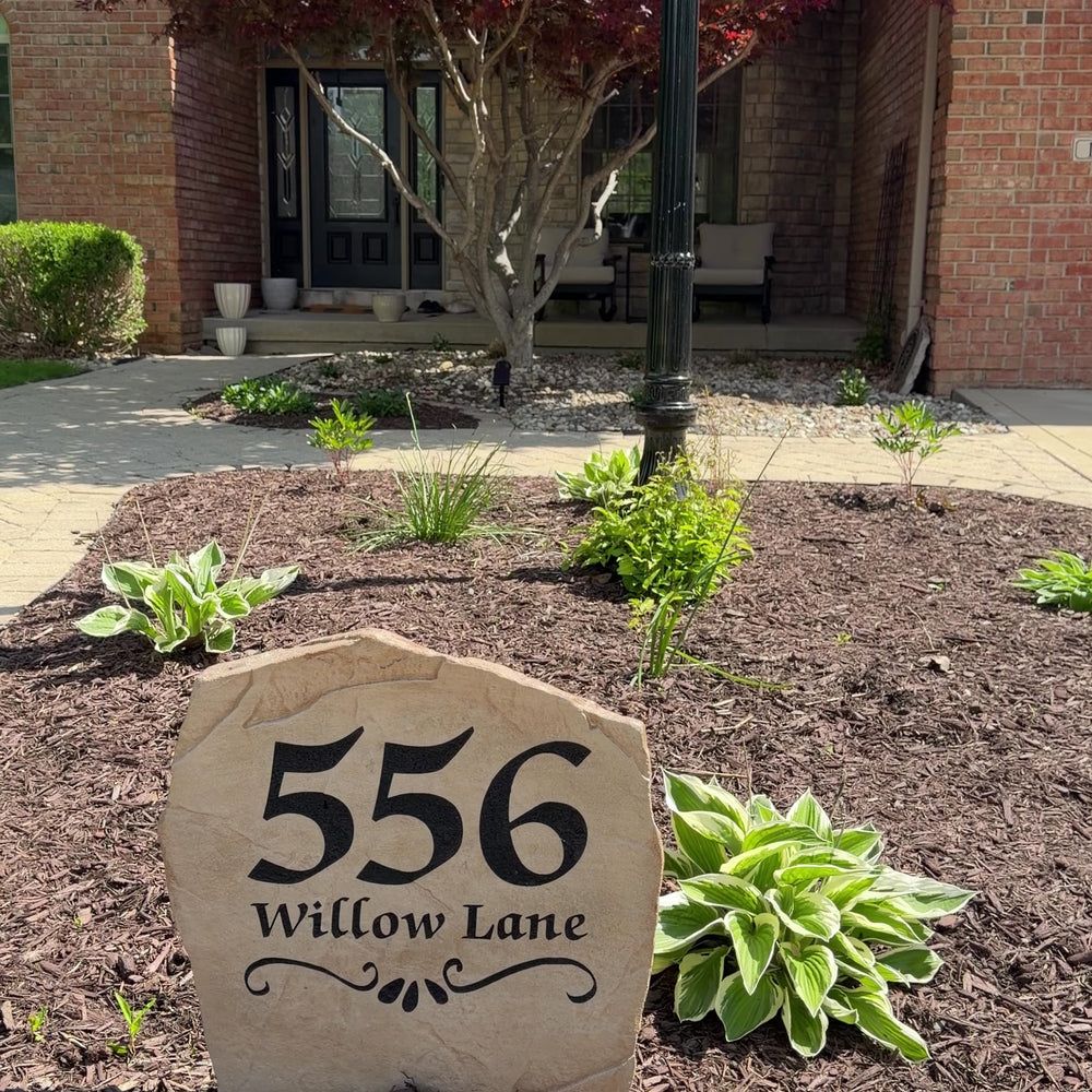 Custom Scroll Address Stone displayed in a landscaped flower bed, featuring personalized house numbers and decorative scroll accents. The durable engraved design provides a timeless address marker for driveways, gardens, and home entrances.